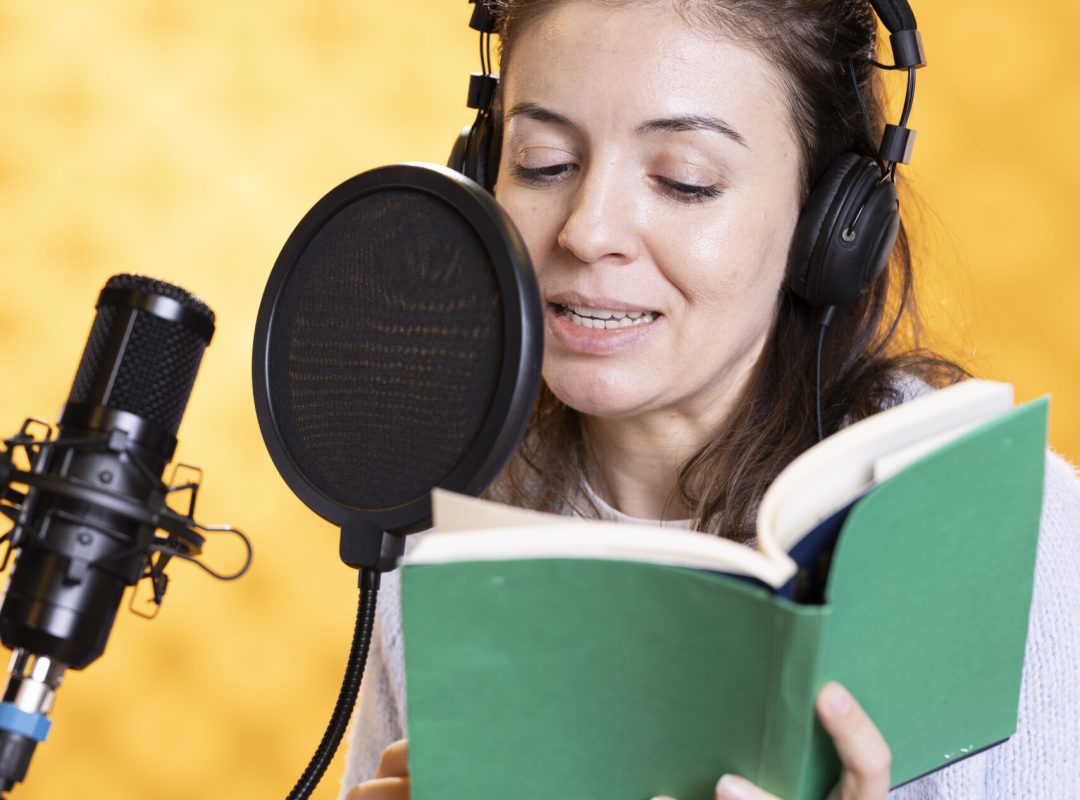 Smiling narrator wearing headphones reading aloud from book into microphone against yellow background. Cheerful person recording audiobook, creating engaging media content for listeners